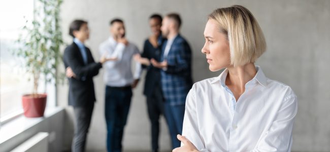 Male Coworkers Whispering Behind Back Of Unhappy Businesswoman In Office